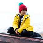 A boy in a Puddleflex Waterproof Fleece Lined Jacket in yellow, sitting on a boat wearing a red hat with a blue pom pom.